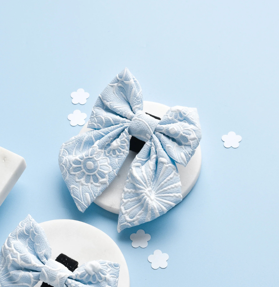 Cocopup London Baby Blue Flower LUXE Sailor Bow Tie on a blue background with decorative paper flowers
