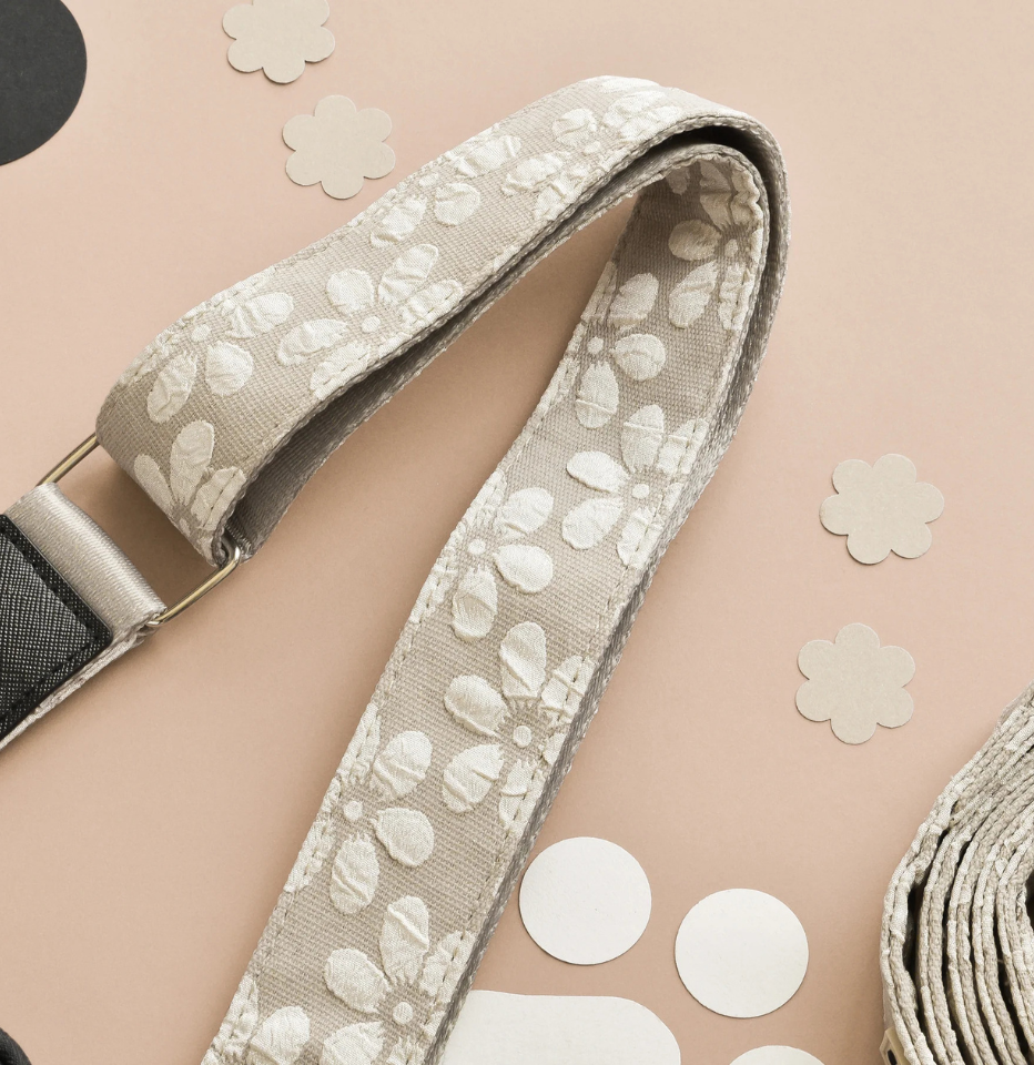 A close-up of a Cocopup London Mocha Flower LUXE Walking Bag Strap, laid diagonally on a peach-colored background with decorative paper cutouts.