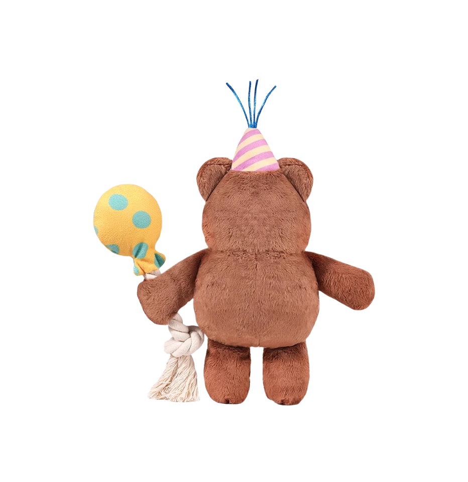 The back of a Hugsmart Yappy Birthday Party Bear Dog Toy, wearing a pink and yellow striped party hat and holding a yellow balloon with blue polka dots and a cream-colored rope tassel, isolated on a white background.