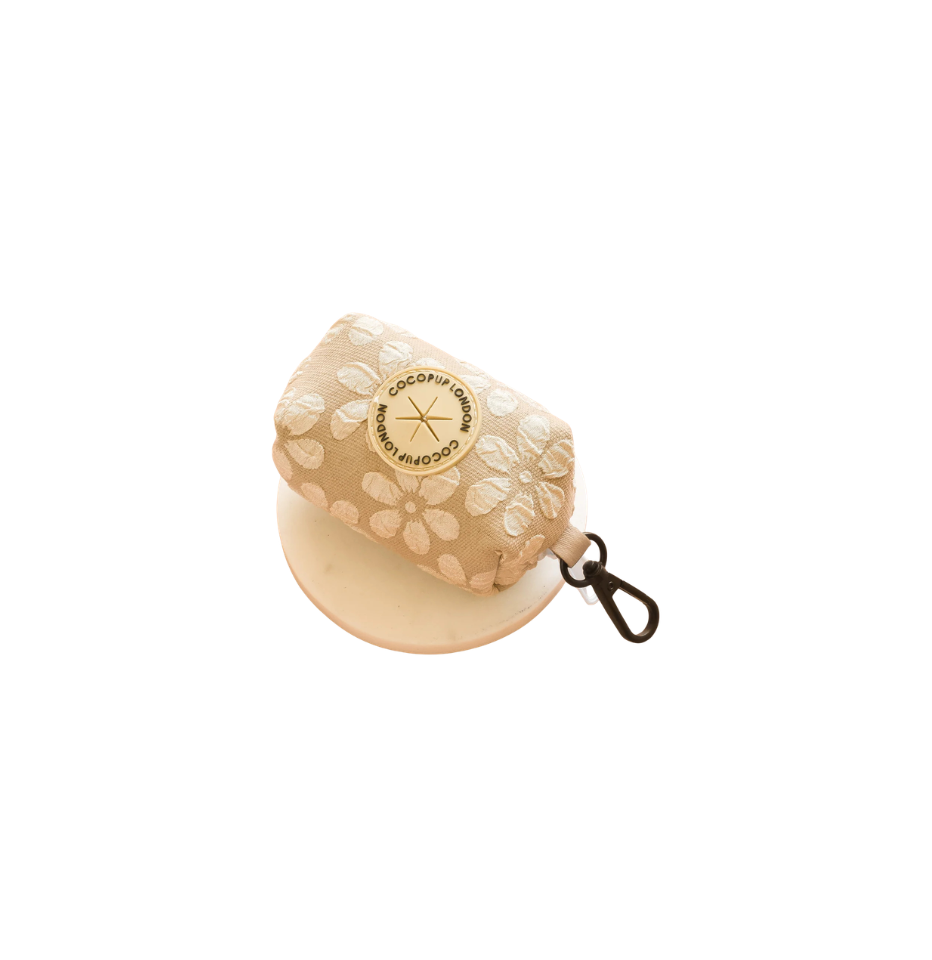 A beige and white floral patterned dog poop bag holder with a circular gold logo reading 'cocopup london' with a black clip and on a white background .