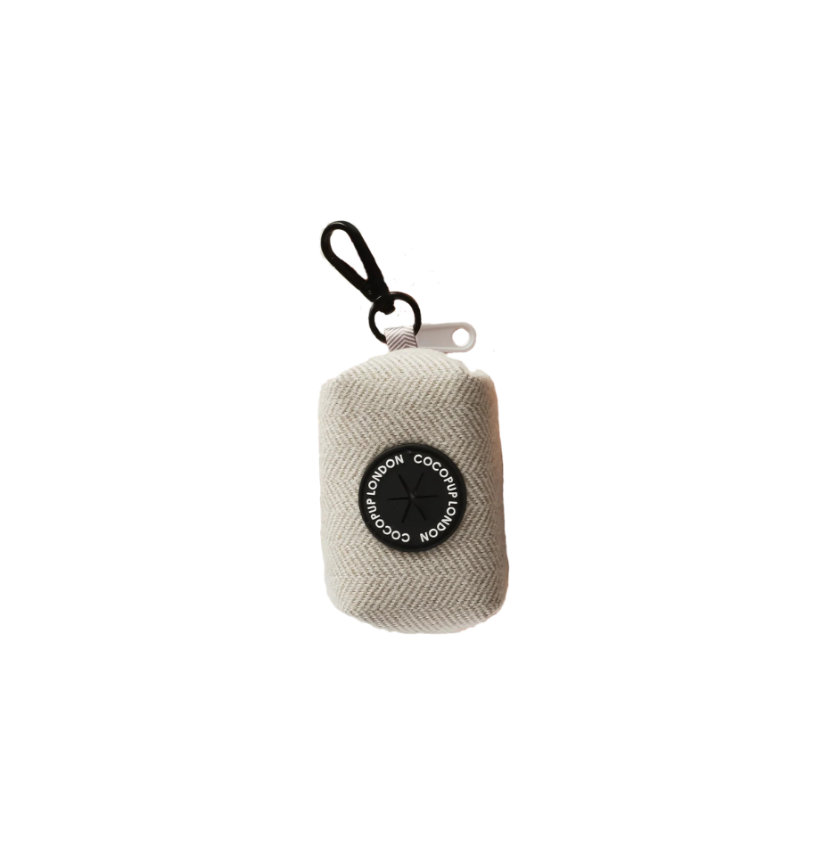 A small light brown herringbone fabric dog poop bag holder with a black circular logo patch and a black carabiner clip, shown on a plain white background.