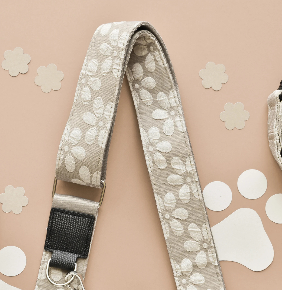 A close-up of a fCocopup London Mocha Flower LUXE Walking Bag Strap, featuring a black plastic buckle and silver metal hardware, laid out on a peach-colored background with decorative paper cutouts.