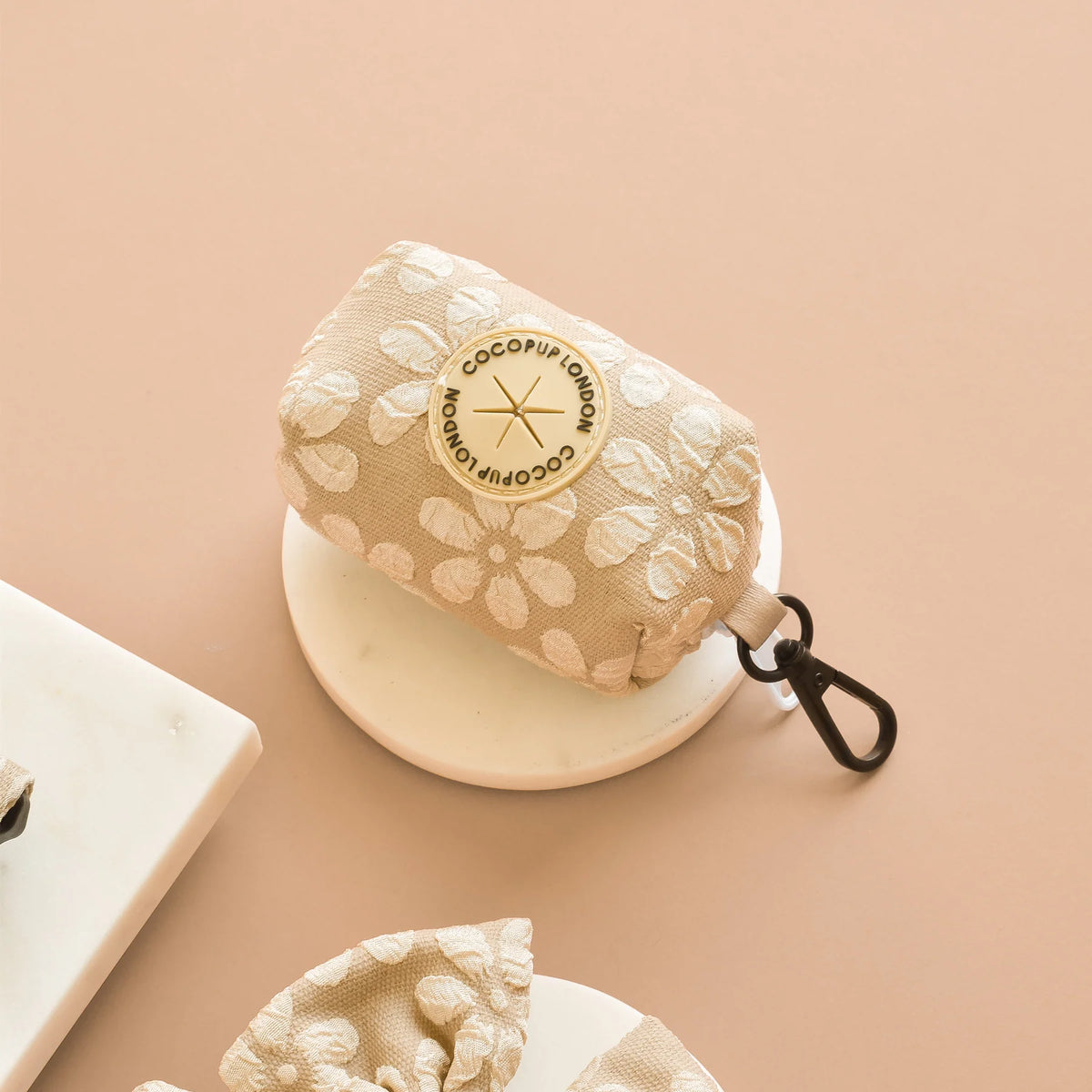 A beige and white floral patterned dog poop bag holder with a circular beige logo reading 'cocopup london' on a peach-pink background.
