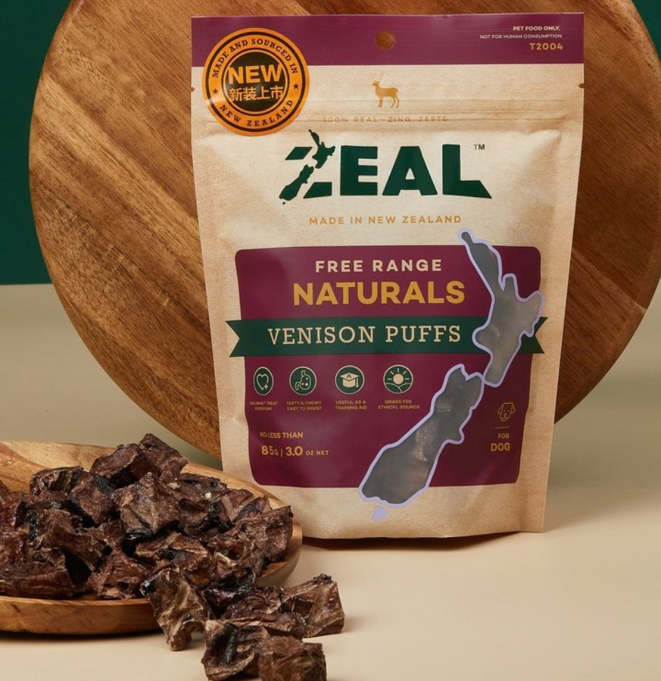 A bag of Zeal Free Range Naturals Venison Puffs - 85g is displayed on a beige surface with a wooden board behind it. The treats are also scattered on a wooden plate in the foreground.